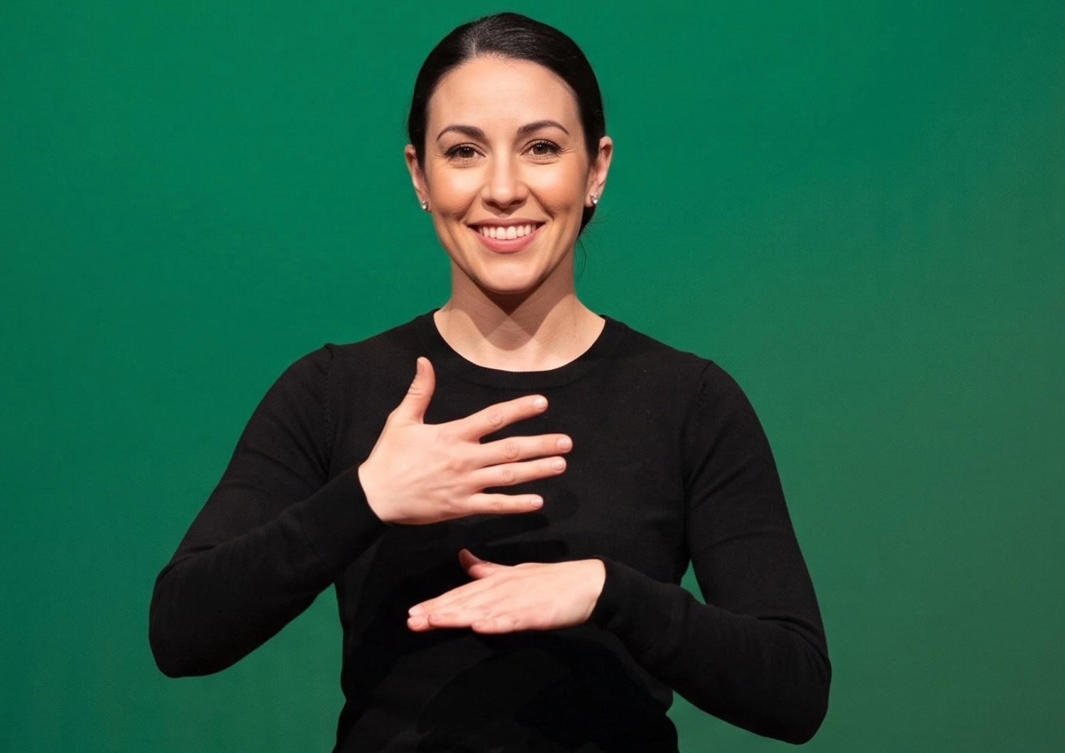 An interpreter in a black long-sleeved top signing in front of an evenly lit, plain green backdrop. She is framed from the waist up with clear space around her hands.